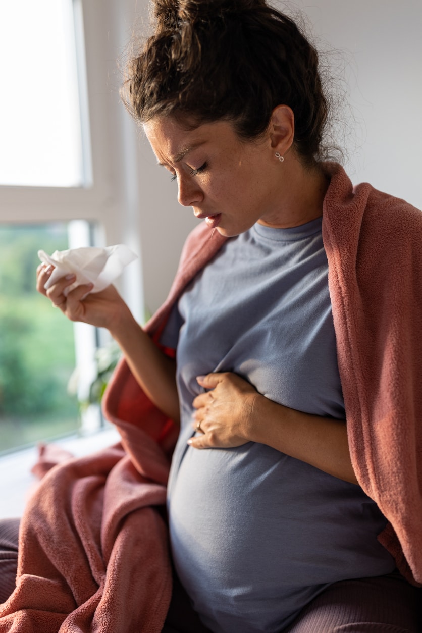 Pregnant woman sitting on bed covered in blanket and sneezing into tissue.